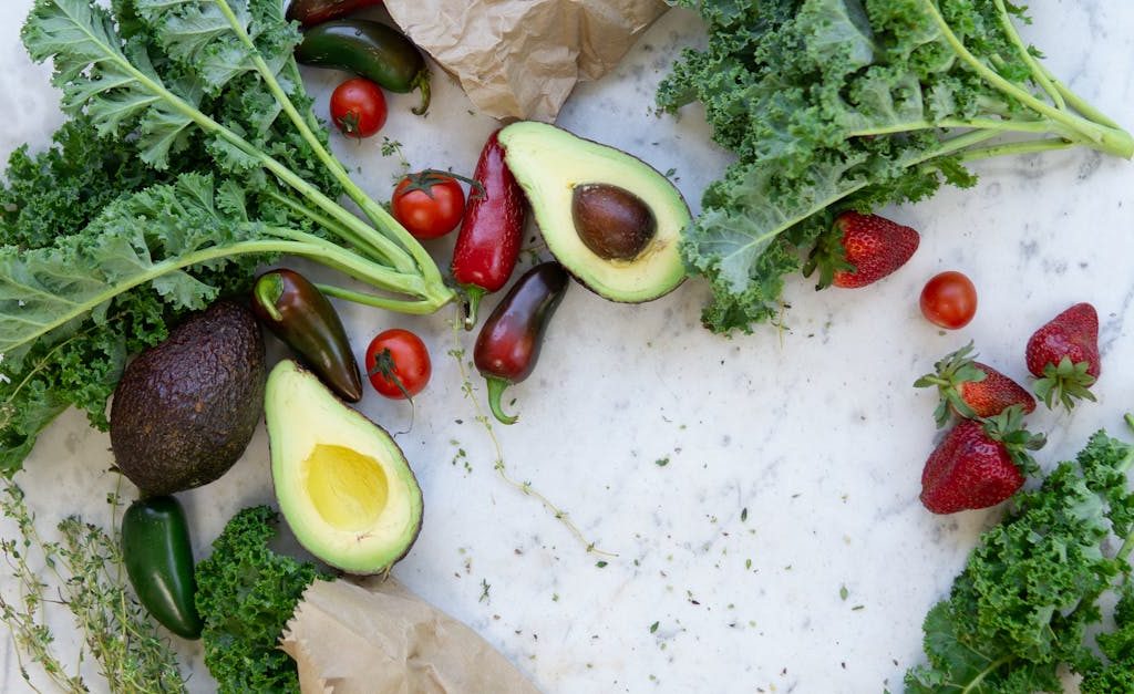 Top view of fresh avocados, kale, tomatoes, and peppers arranged on a marble surface.